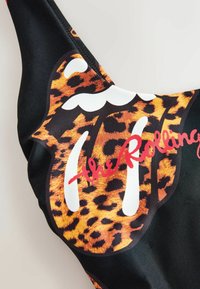 Közeli kép fekete anyagról, amelyen a Rolling Stones nyelv logója látható leopárdmintával, és piros kézírásos szöveg van ráírva.