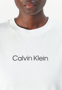 Camiseta de algodón blanca con escote de cuello redondo y logo negro de "Calvin Klein" en el pecho. Textura suave y corte relajado.