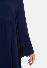 Mörkblå klänning med lös passform, lång ärm, vecken nära fållen och sidoklyftor för extra rörelse. Mjukt, lättviktigt tyg.