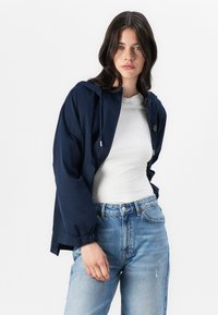Marineblå lett jakke med hette, elastiske mansjetter og glidelås foran. Sammen med lys blå høyt liv jeans.