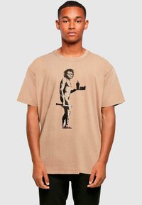 Beige Baumwoll-T-Shirt mit einem Grafikmotiv eines Steinzeitmenschen, der ein Tablett mit Essen und einem Getränk hält, mit Rundhalsausschnitt und lockerer Passform.