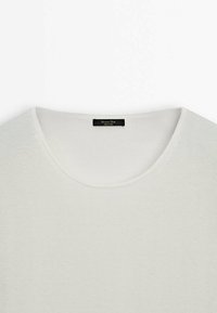 Pull en maille blanche douce avec un col rond présenté à plat, montrant la texture et une étiquette de marque noire au col.
