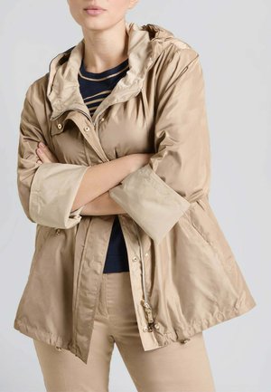 Persona con le braccia incrociate che indossa una giacca beige con cappuccio sopra una maglia blu navy e pantaloni beige su uno sfondo semplice.