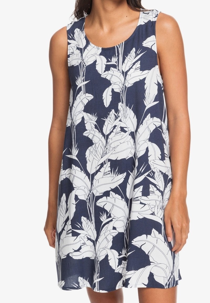 Robe sans manches bleu marine avec un motif de feuilles tropicales blanches portée par une personne, longueur genou et coupe ample.