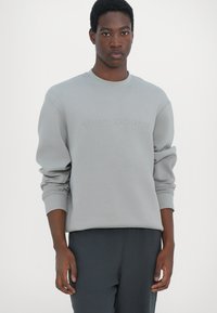 Light grey sweatshirt featuring ribbed cuffs and neckline, with 'Armani Exchange' embossed on the front. Worn with dark trousers.