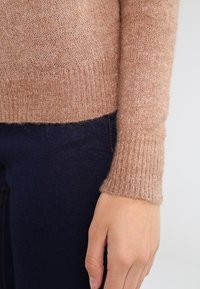 Jasnobrązowy sweter z dzianiny o miękkiej fakturze, z prążkowanymi mankietami i dopasowanym krojem, w połączeniu z ciemnoniebieskimi dżinsami.