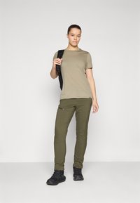 Pantalones ligeros verde oliva con cremalleras, combinados con una camisa de manga corta beige. El modelo lleva una mochila negra. Tejido texturizado.