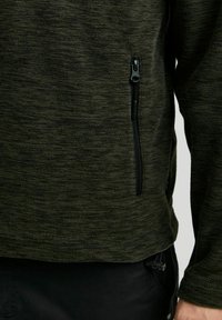 Fleece jack in donkergroen met een gestructureerd patroon, voorzien van een ritssluiting aan de linkerkant en zwarte hardware-accenten.