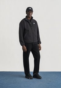 Νέος άνδρας με μαύρο φλις μπουφάν, μαύρο παντελόνι, μαύρα αθλητικά παπούτσια και μαύρο καπέλο με λευκό λογότυπο, στέκεται σε μπλε χαλί.