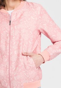 Różowa kurtka bomberka z białym kwiatowym wzorem, ściągacze przy kołnierzu i mankietach, zamek błyskawiczny i kieszenie boczne. Miękka tkanina o gładkiej fakturze.