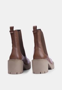 Botas de cuero marrón hasta el tobillo con paneles laterales elásticos, tacones gruesos beige y un acabado suave. Consta de tiradores para ponerlas y quitarlas fácilmente.