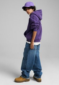 Fioletowa bluza z kapturem, biała koszulka pod spodem, luźne niebieskie dżinsy, beżowe buty robocze i dopasowana fioletowa czapka.