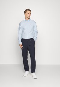 Camicia azzurra chiara con maniche lunghe, abbinata a pantaloni blu navy e sneakers bianche. La camicia presenta un piccolo logo sul petto.