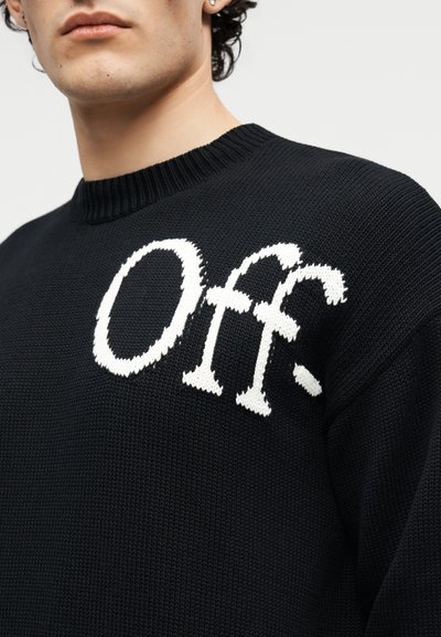 Κοντινή λήψη ενός ατόμου που φοράει μαύρο πλεκτό πουλόβερ με μεγάλα λευκά γράμματα που γράφουν "Off" στο στήθος.