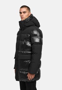 Svart puffjacka med blank yta, justerbar huva, ficka med dragkedja på bröstet och sidofickor, som har en strömlinjeformad, vadderad design.