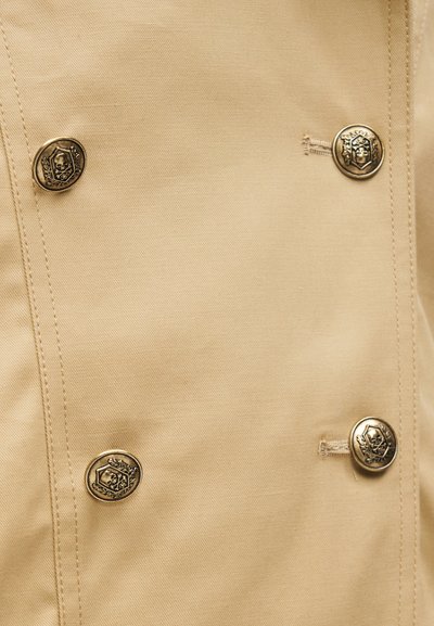 Μπεζ ύφασμα με τέσσερα διακοσμητικά μεταλλικά κουμπιά που απεικονίζουν σχέδια κρανίου και δάφνινου στεφάνου, τοποθετημένα σε ορθογώνιο μοτίβο.