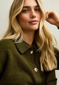 Blond kvinna med löst hår bär en grön stickad tröja med stora knappar, rör vid håret nära örat, neutral bakgrund.