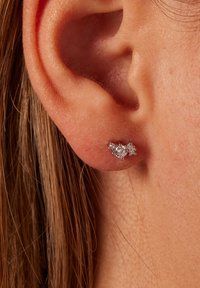 Pendientes de plata con tres piedras preciosas transparentes dispuestas en un racimo, sobre un tono de piel natural con cabello lacio al fondo.
