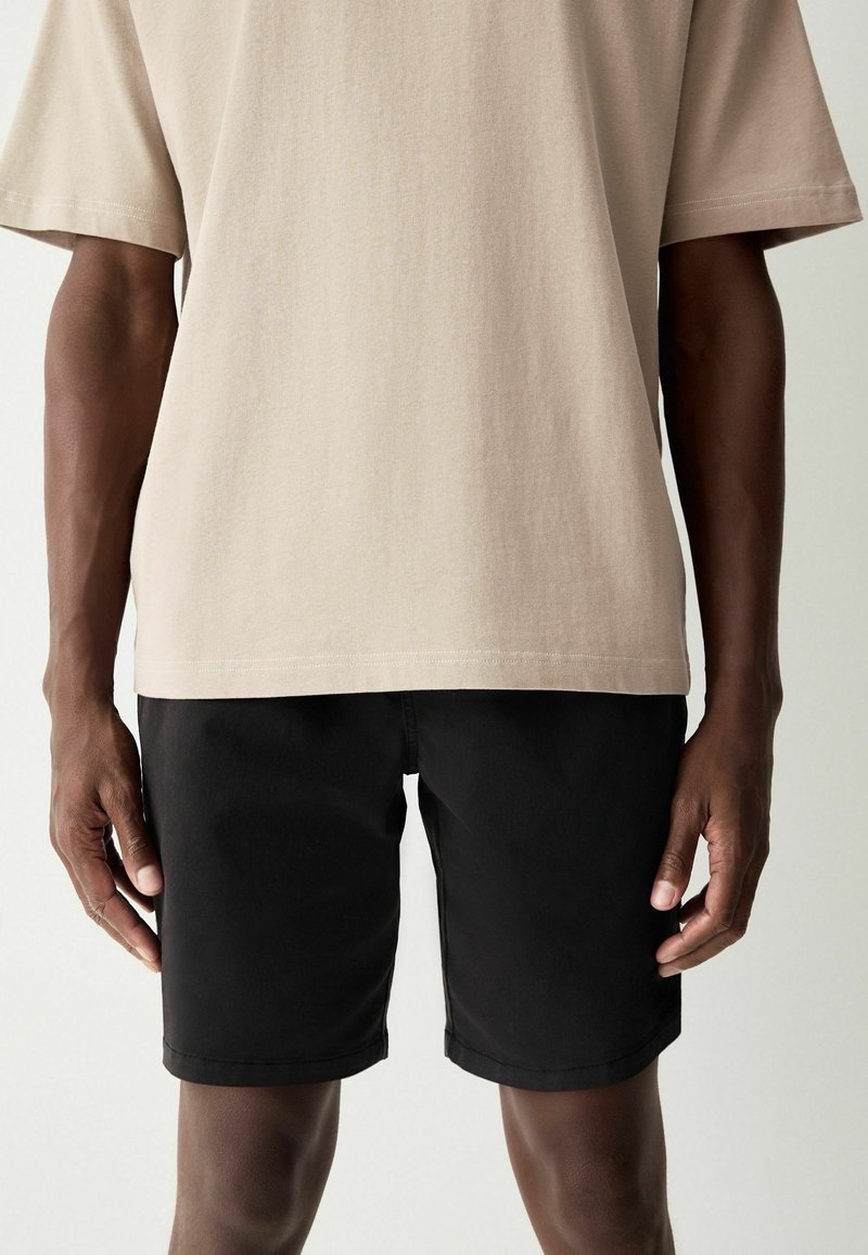 Camicetta beige a maniche corte con una finitura testurizzata abbinata a pantaloni corti neri. I pantaloni corti hanno un taglio dritto e dei risvolti.