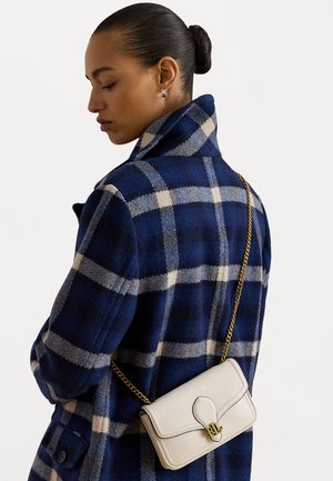 Ženska s počešljano nazaj pričesko nosi plašč v mornarsko modri in beli karo vzoru, v rokah drži majhno belo usnjeno torbico z zlatim verižnim ročajem in RL logotipom.