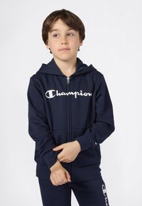 Sudadera con cremallera en color navy con logo blanco de "Champion", fabricada en una mezcla de algodón, con bolsillos frontales y capucha ajustable. Modelo luciendo pantalones a juego.