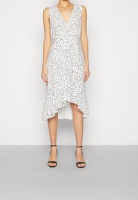 Robe blanche sans manches avec des pois noirs, décolleté en V et ourlet asymétrique. Tissu léger avec une taille cintrée, complétée par des talons noirs.