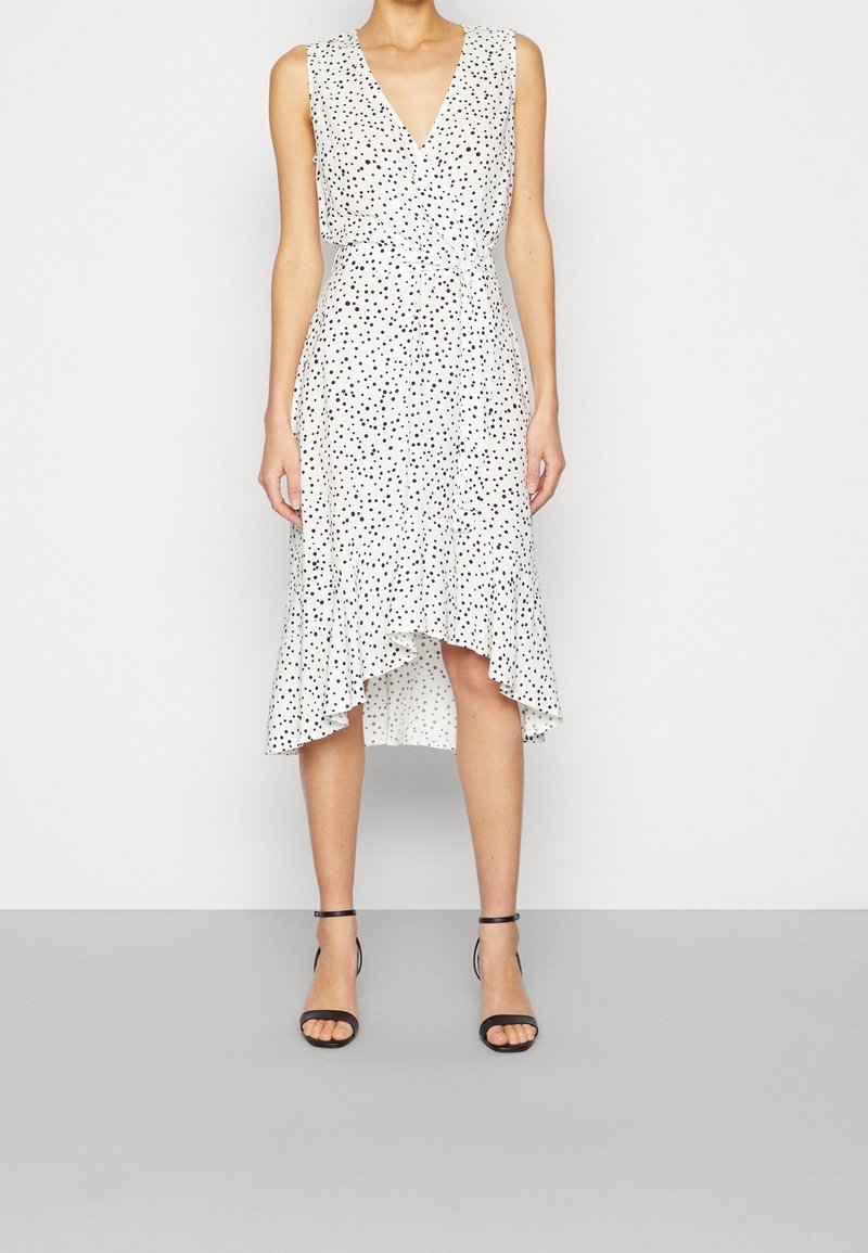 Robe blanche sans manches avec des pois noirs, décolleté en V et ourlet asymétrique. Tissu léger avec une taille cintrée, complétée par des talons noirs.