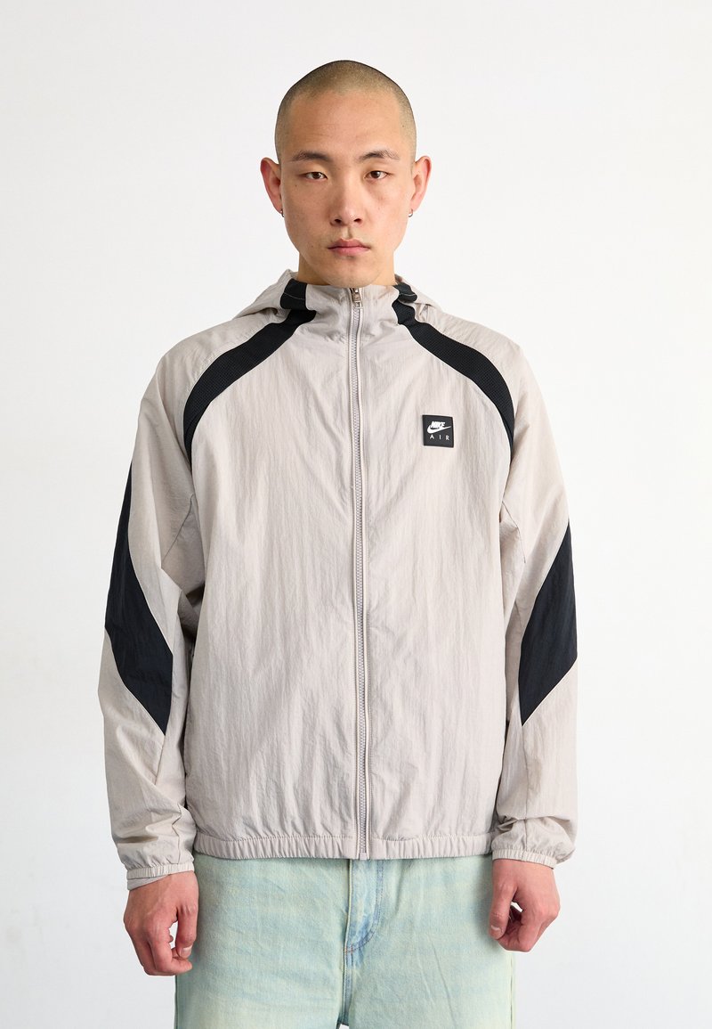 Light gray windbreaker jacket with black accents, featuring a front zipper, hood, and elastic cuffs. Nike Air logo on the chest.