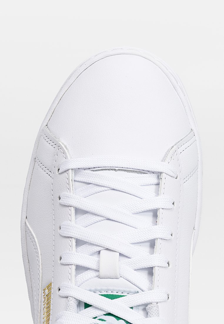 Weißer Sneaker mit oberer Glattlederfläche, flachen Schnürsenkeln und grüner Innensohle. Auffällige goldene Logo-Akzente an der Seite.