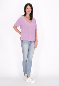 Világoslila, bordázott kötésű, rövid ujjú cardigan, V-nyakkal és gombos zárással, világoskék skinny farmernadrággal és fehér sportcipőkkel párosítva.