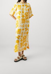 Robe imprimée jaune à manches courtes, présentant des motifs floraux abstraits et géométriques sur un fond blanc. Portée avec des sandales blanches.