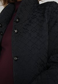 Sort dunjakke med et tekstureret diamantmønster, der har en høj krave og flere sorte knapper til lukning.