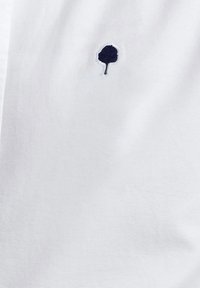 Tejido de algodón blanco con un pequeño logo de árbol bordado en color navy. La textura es suave con un brillo sutil.