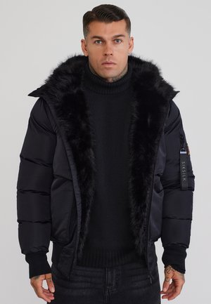 Uomo che indossa una giacca imbottita nera con cappuccio foderato in pelliccia e maglione nero a collo alto, in piedi contro uno sfondo chiaro e uniforme.