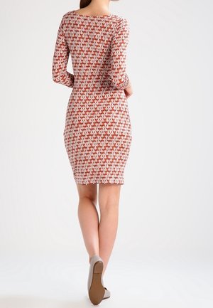 Mujer de pie de espaldas a la cámara, vistiendo un vestido de manga larga hasta la rodilla con un patrón geométrico en rojo y blanco, y zapatos beige.