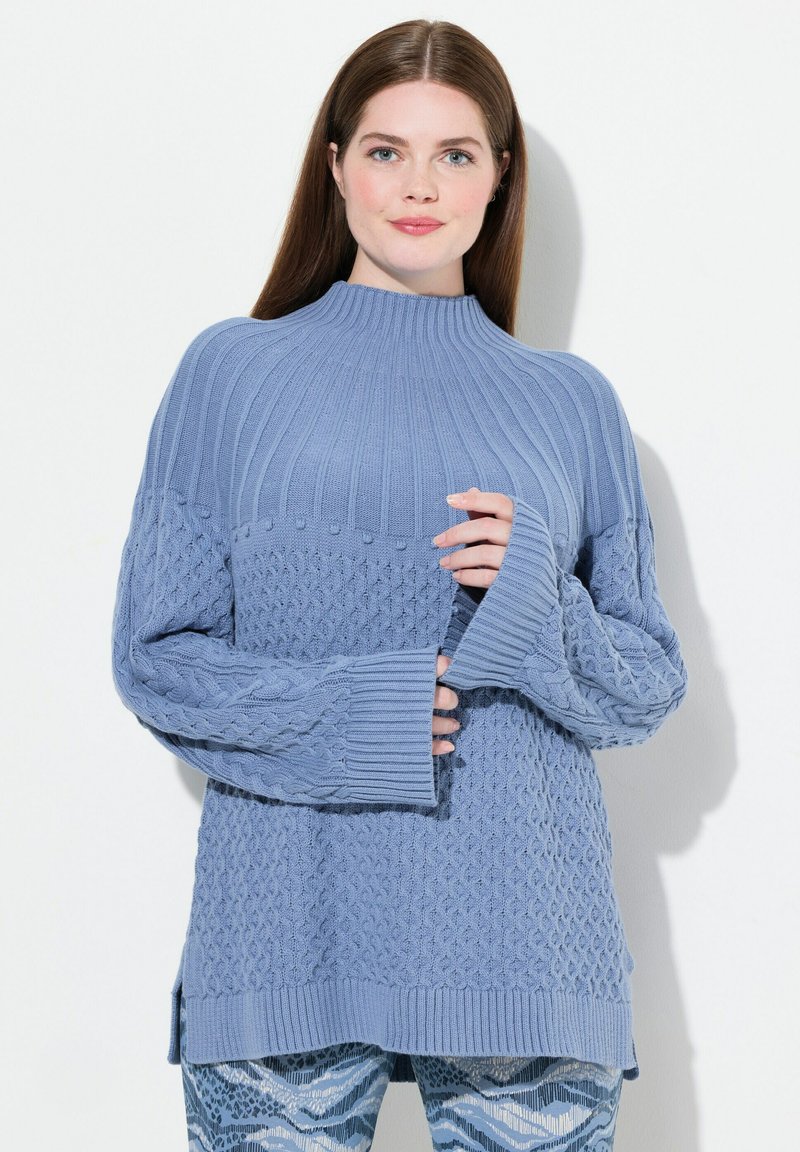 Ljusblå stickad tröja med ribb- och texturmönster, hög krage och långa ärmar, med sidoöppningar och en avslappnad passform.