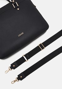 Musta teksturoidusta materiaalista valmistettu käsilaukku, jossa on kullanväriset metalliosat, logo ja irrotettava musta olkahihna, jossa on kohopainettu brändi. Sileä pinta ja jämäkkä muoto.