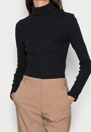 Personne portant un pull à col roulé noir tacheté et un pantalon beige taille haute, les mains dans les poches sur fond blanc.