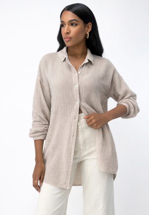 Frau mit langen schwarzen Haaren, die eine beige strukturierte Hemdbluse und eine weiße Hose trägt, steht vor einem schlichten weißen Hintergrund.