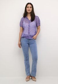 Blusa corta a maniche corte di colore viola con bottoni, abbinata a jeans a zampa d'elefante di colore blu chiaro e scarpe con tacco beige. Tessuto morbido.