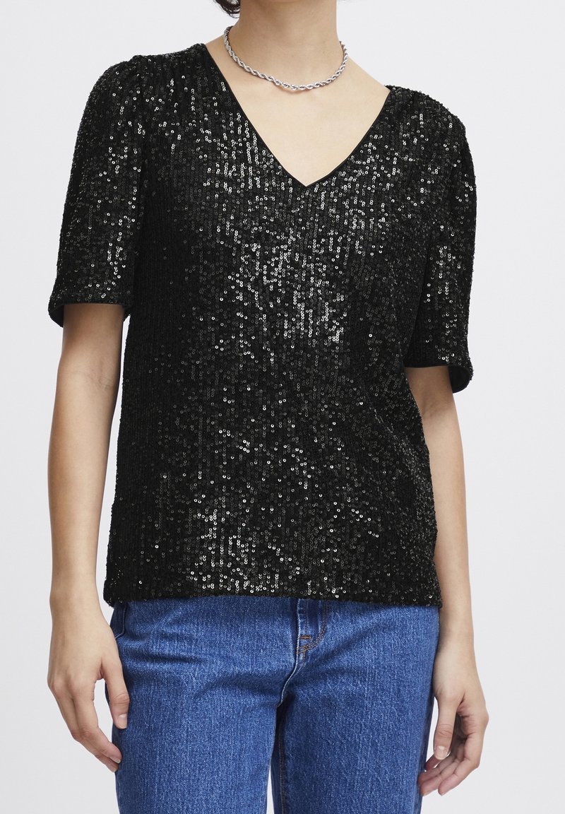 Svart paljett-topp med V-ringning och korta puffärmar, med en texturerad yta. Kombinerad med blå denimjeans.