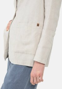 Lichtbeige linnen blazer met een steekzak, rechte snit en opgerolde mouwen. Verfraaid met een klein bruin merktekentje.