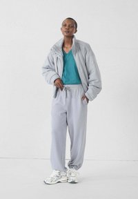 Lichtgrijze oversized jas, teal v-hals t-shirt, lichtgrijze sweatpants en zilveren sportschoenen. Glad fabric, ontspannen pasvorm.