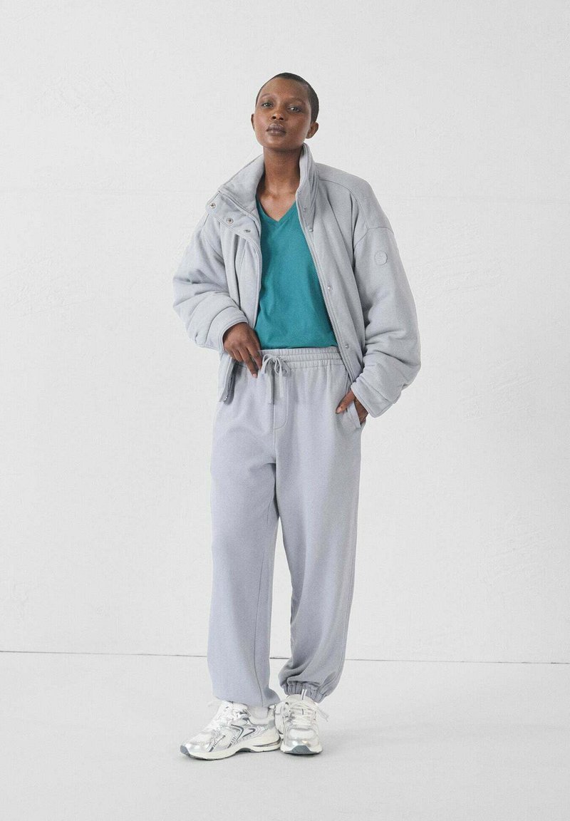 Lichtgrijze oversized jas, teal v-hals t-shirt, lichtgrijze sweatpants en zilveren sportschoenen. Glad fabric, ontspannen pasvorm.