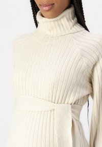 Pulover ribbed de culoare crem, cu un guler înalt și un detaliu cu curea la talie. Textură moale, tricotată, cu linii verticale subtile.