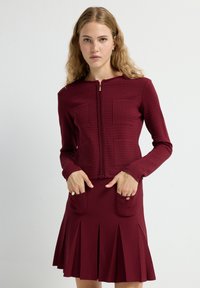 Chaqueta de cremallera color marrón burdeos con un patrón de rejilla texturizada y dos bolsillos frontales, combinada con una falda acampanada a juego con pliegues.