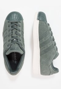 Zapatillas de ante gris con una puntera de goma, cordones, detalles perforados y una entresuela blanca contrastante. Diseño clásico de tres rayas.