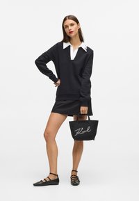Mujer con una camisa negra de manga larga y cuello blanco, falda negra y bailarinas negras, sosteniendo un bolso negro con la palabra "Karl" escrita en él.