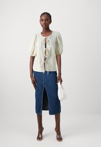 Blusa gialla chiara con maniche a sbuffo e dettaglio annodato davanti, abbinata a una gonna midi in denim scuro con spacco anteriore. Tacchi trasparenti e borsa bianca.