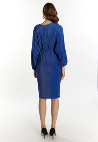 Mėlynas blizgučių suknelė su ilgomis, plačiomis rankovėmis ir įvyniuotu liemeniu. Kelio ilgio dizainas pasižymi lygia tekstūra ir subtiliu žvilgesiu.